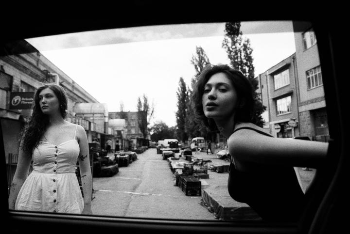 Two women are visible through a car window, one standing on the street and the other leaning out of the car, with a street market in the background.