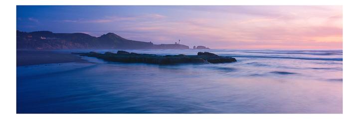 A long exposure captures the movement of waves on a beach with a lighthouse and mountains in the distance at sunset.