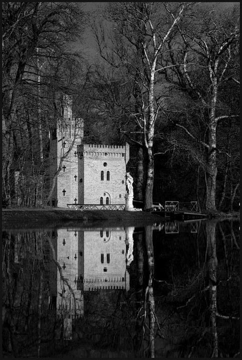 A small castle with a statue in front of it is reflected in a body of water, surrounded by bare trees.