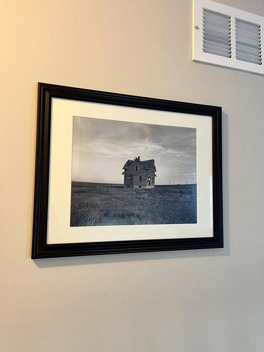 A framed black and white photograph of an abandoned house in a field hangs on a wall next to a vent.
