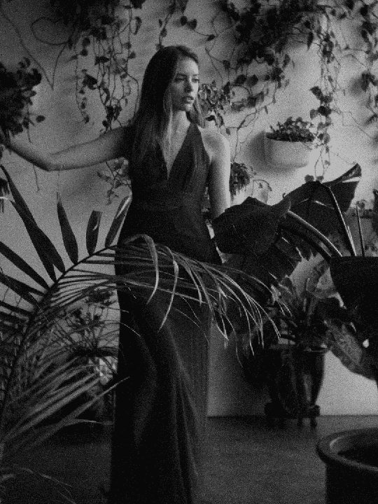 A woman in a long dress stands among many plants in a greenhouse.