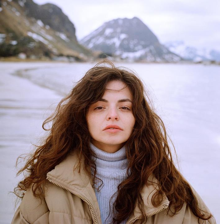 A woman with long, curly hair and a puffer jacket stands in a snowy landscape with mountains in the background.