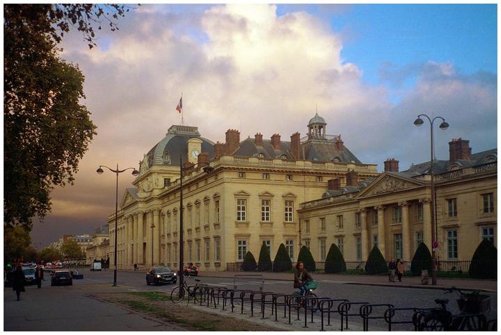 A large classical building with a domed roof and a French flag on top stands next to a street with cars and people.