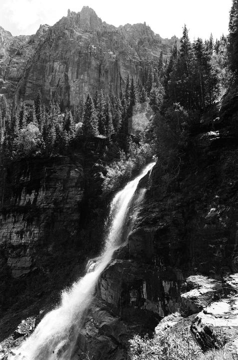 A waterfall cascades down a rocky cliff face surrounded by pine trees and mountains.