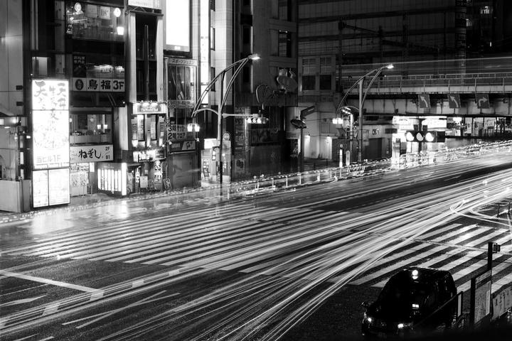 A black car is parked on a city street at night with light trails from traffic and illuminated buildings.