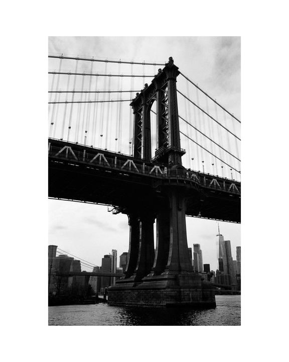 A black and white photograph shows the Manhattan Bridge towering over water with the New York City skyline in the background.