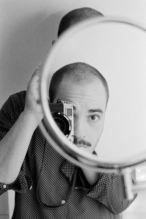 A man with a mustache holds a camera to his eye, reflected in a round mirror.