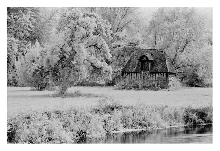 An old wooden house with a dormer window stands next to a river, surrounded by trees and tall grass.