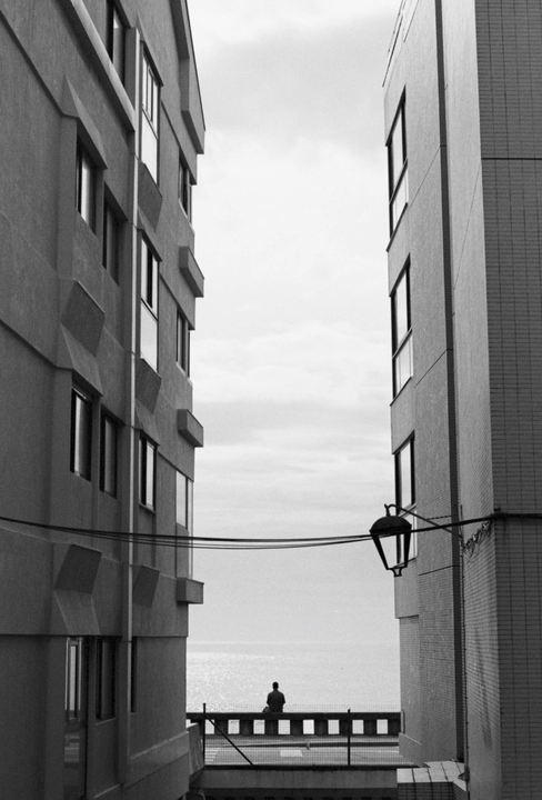 A person sits on a balcony overlooking the ocean, framed by two tall buildings.