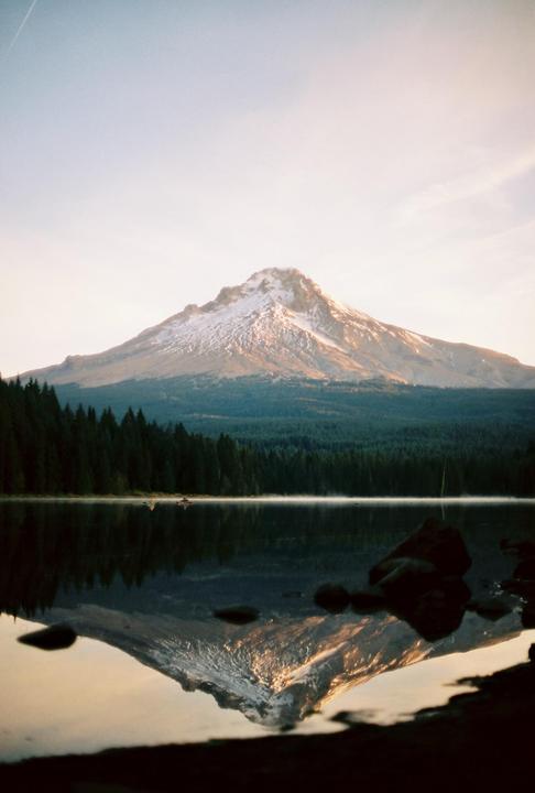 A snow-capped mountain is reflected in a still lake surrounded by a forest.