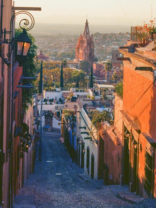 A cobblestone street leads down a hill towards a city with a large church in the distance at sunset.