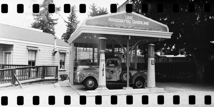 A black and white photograph shows a vintage gas station with a truck parked underneath its canopy.