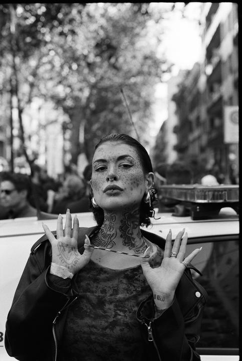 A woman with tattoos on her face, neck, and hands poses with her hands up in front of a vehicle.