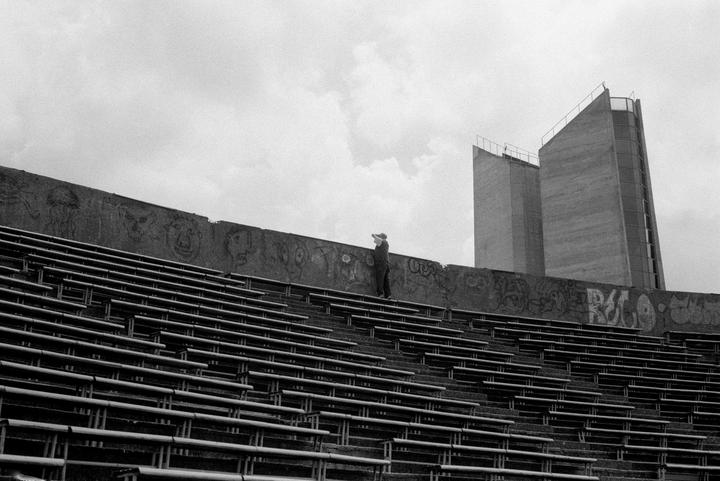 A person stands on a concrete wall with graffiti above rows of empty stadium seating under a cloudy sky.