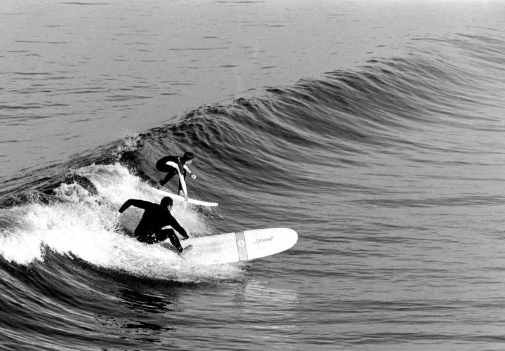 Two surfers ride a wave, one in front of the other, with the ocean stretching out behind them.