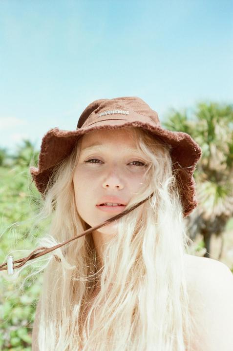 A woman with long blonde hair and a brown bucket hat looks directly at the viewer.