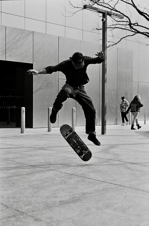 A man on a skateboard is captured mid-air, performing a trick on a paved surface next to a modern building.