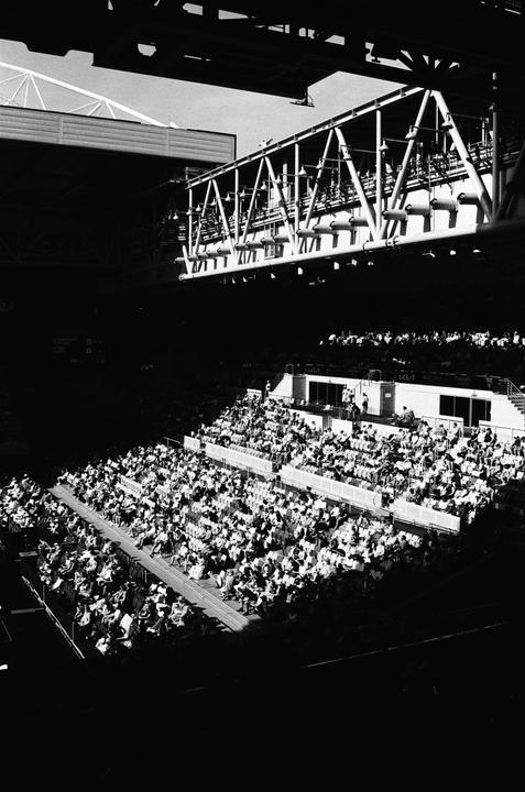 A large crowd of people sits in the stands of a stadium, with a large metal structure overhead.