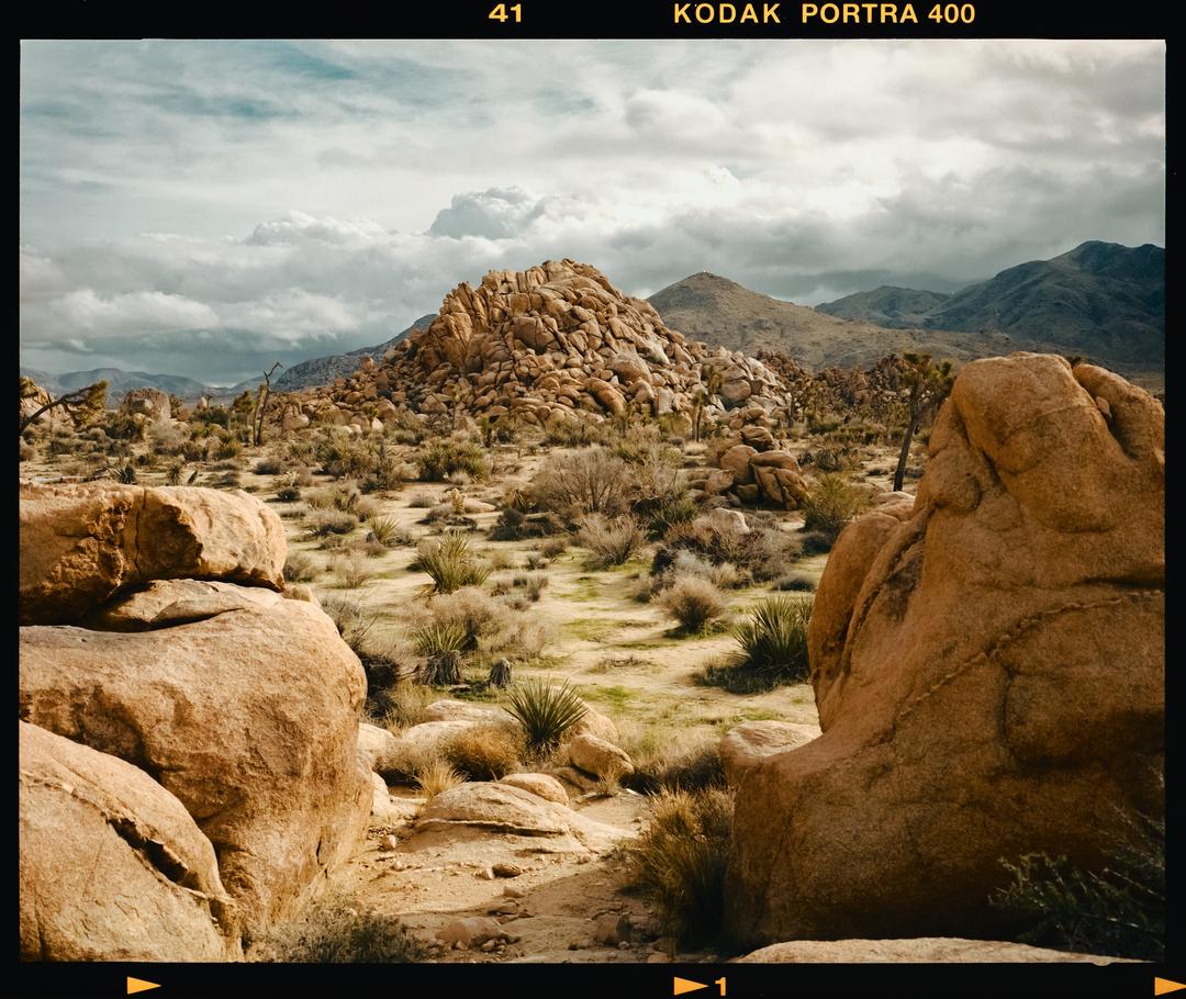 A desert landscape features large rock formations, sparse vegetation, and mountains under a cloudy sky.