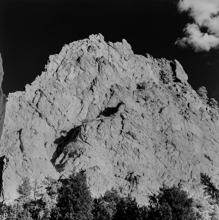A large rock formation rises above a line of trees under a dark sky with a single cloud.