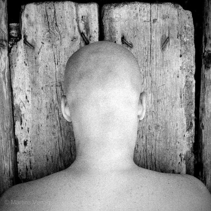 A person with a shaved head is seen from behind, facing a wooden wall with exposed nails.