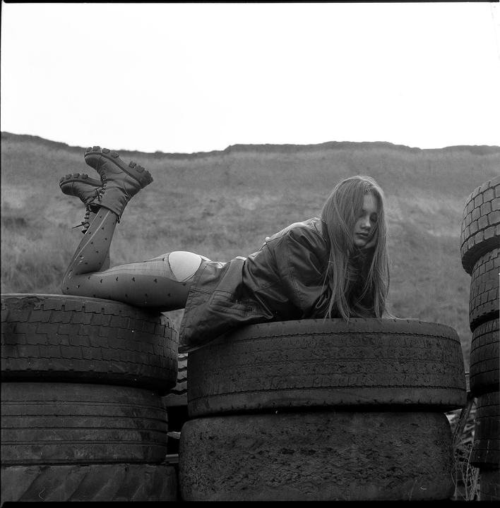 A woman in a leather jacket and patterned tights lies on a stack of tires with a hill in the background.