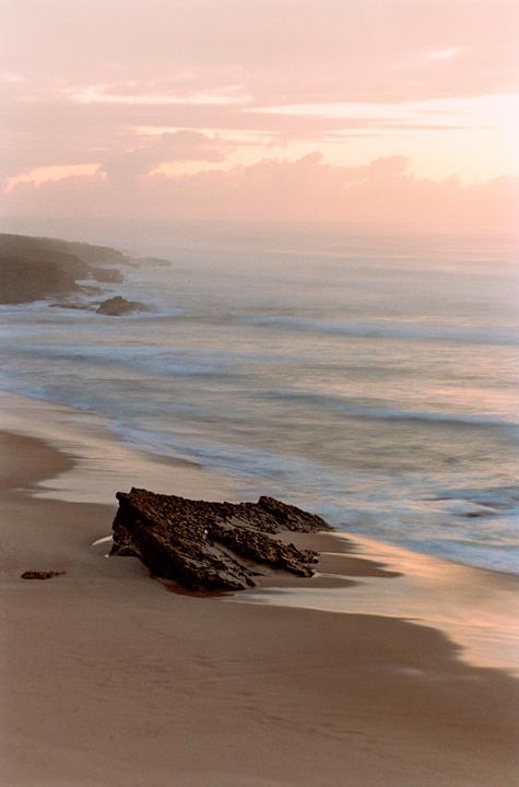 A rocky coastline with waves washing onto a sandy beach is visible under a pastel sky.