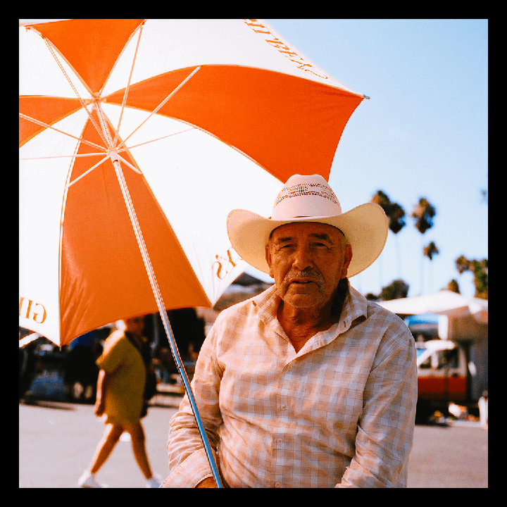 A man wearing a cowboy hat and a plaid shirt sits under a large orange and white umbrella.