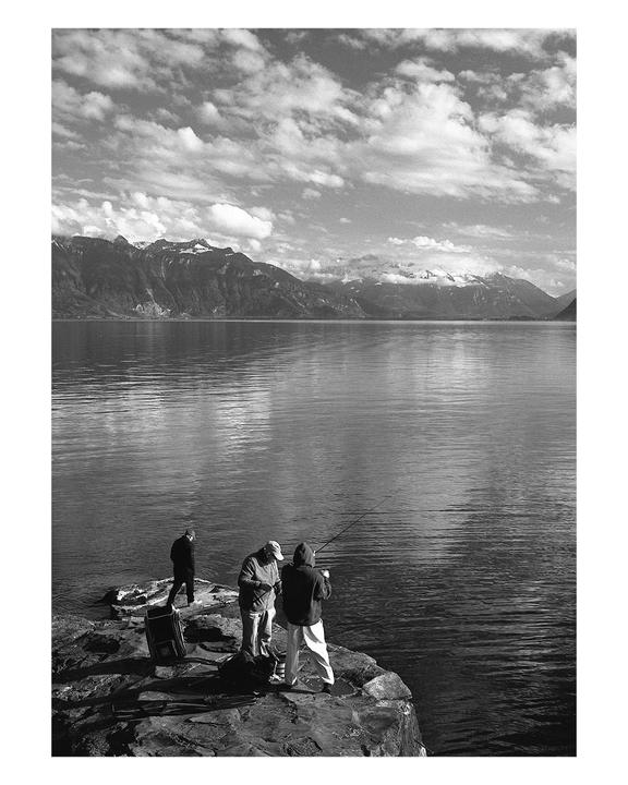 Three men are fishing from a rocky shore with a large lake and mountains in the background under a cloudy sky.
