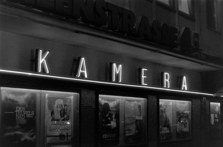 A movie theater marquee with neon letters spelling "KAMERA" is visible above several movie posters.