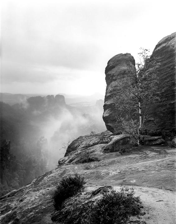 A black and white landscape shows a rocky cliff overlooking a valley filled with fog and distant rock formations.