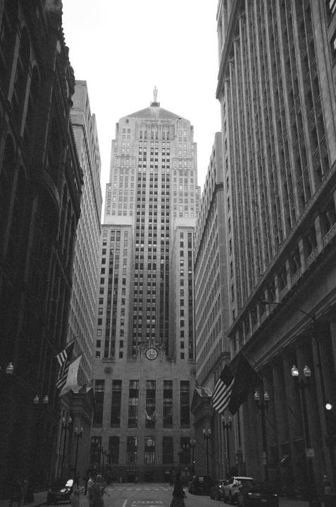 A tall building with a clock and flags on its facade stands at the end of a street lined with other buildings.