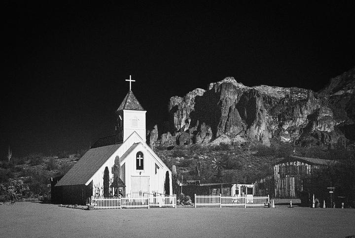 A white church with a steeple and a cross stands in front of a large mountain.