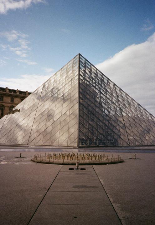A glass pyramid stands under a blue sky with clouds, with a circular fountain in the foreground.