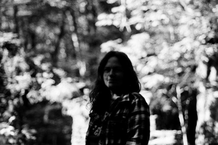 A woman with long hair and a plaid shirt stands in a forest with bright, out-of-focus foliage in the background.