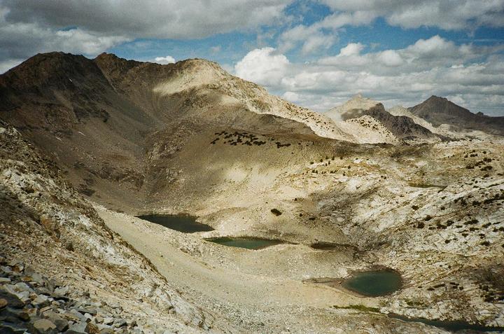 A rugged mountain landscape features several small lakes nestled among rocky terrain under a cloudy sky.