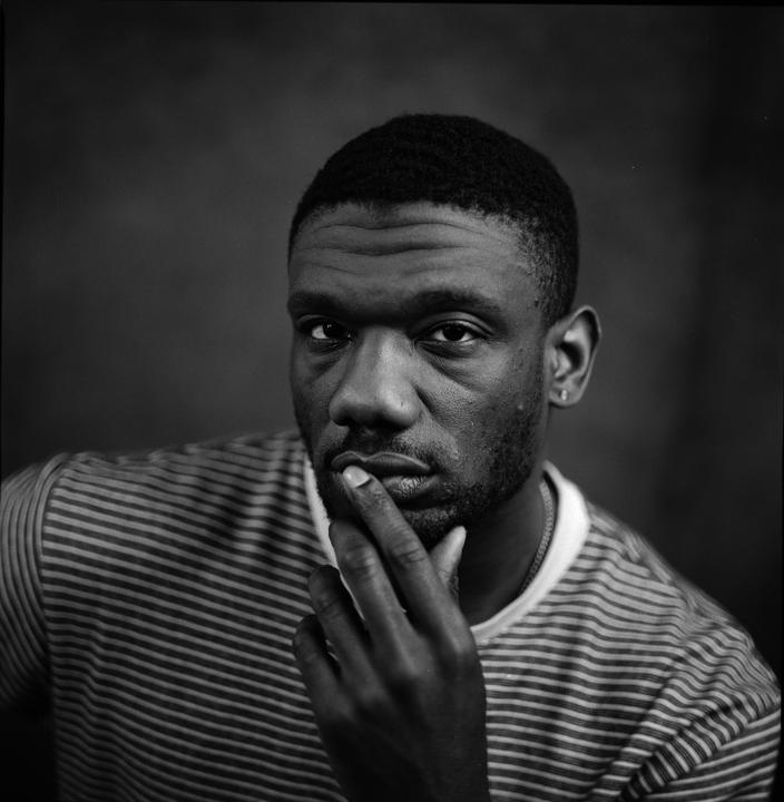 A man with a striped shirt and a beard looks at the camera with his finger on his lips.