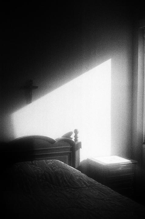 Sunlight streams into a dark bedroom, illuminating a bed and a nightstand.