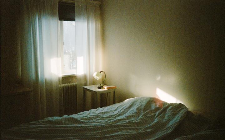 Sunlight streams through a window with sheer curtains into a bedroom with a bed and a nightstand with a lamp and a book.