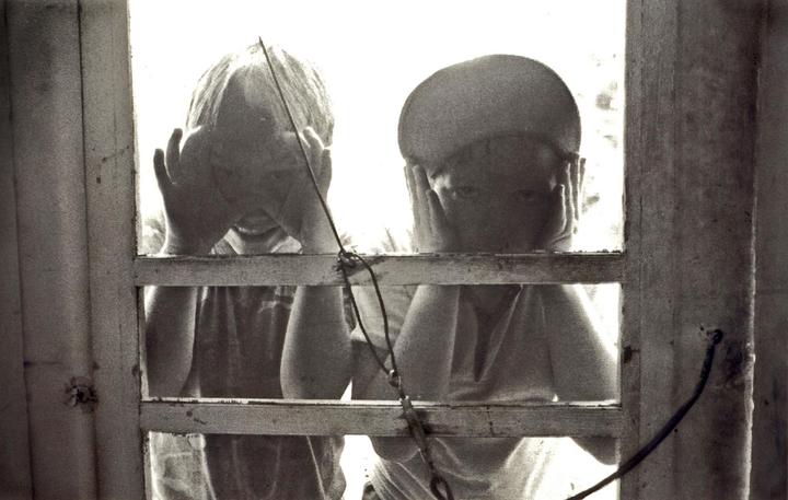 Two children are looking through a screen door, one with hands covering their face and the other with hands on their cheeks.