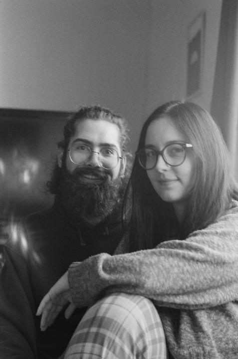 A man with a beard and a woman with long hair sit close together, both wearing glasses and looking at the viewer.
