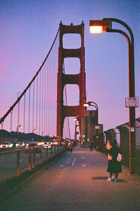A couple stands on the Golden Gate Bridge at dusk, with cars driving past and the city lights in the distance.