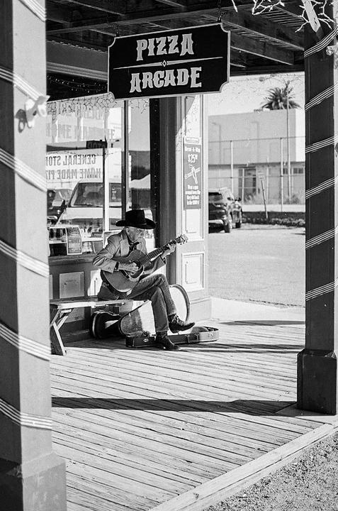 A man wearing a cowboy hat sits on a bench and plays a guitar in front of a pizza arcade.