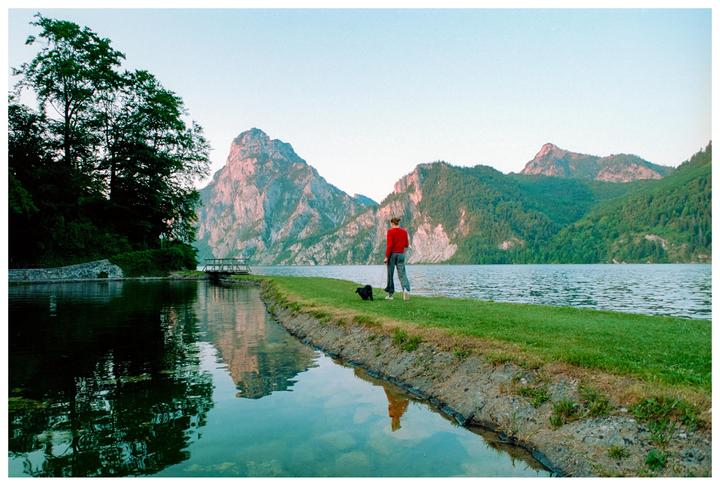 A person and a dog stand on a grassy bank next to a lake with mountains in the background.
