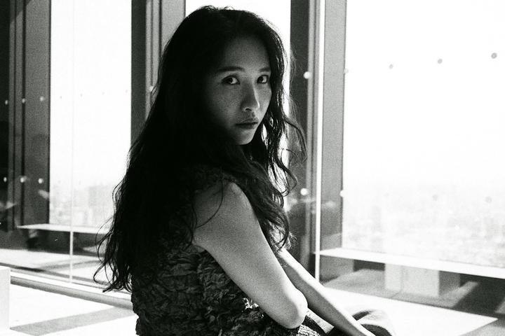 A woman with long dark hair looks over her shoulder at the viewer, sitting in front of a large window.