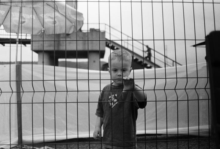 A young boy stands behind a metal fence, looking through the wires.