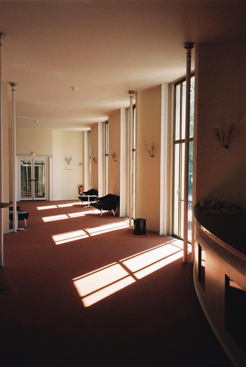 Sunlight streams through tall windows onto a red carpeted hallway with chairs and tables.