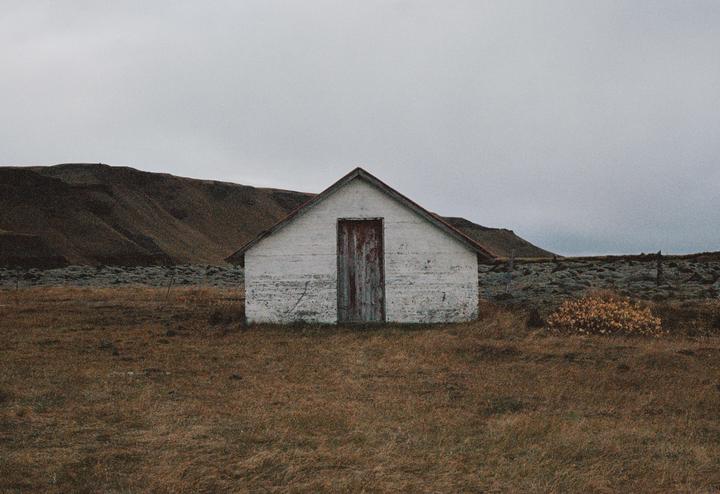 A small white shed with a red door stands in a field with a mountain in the background under a cloudy sky.