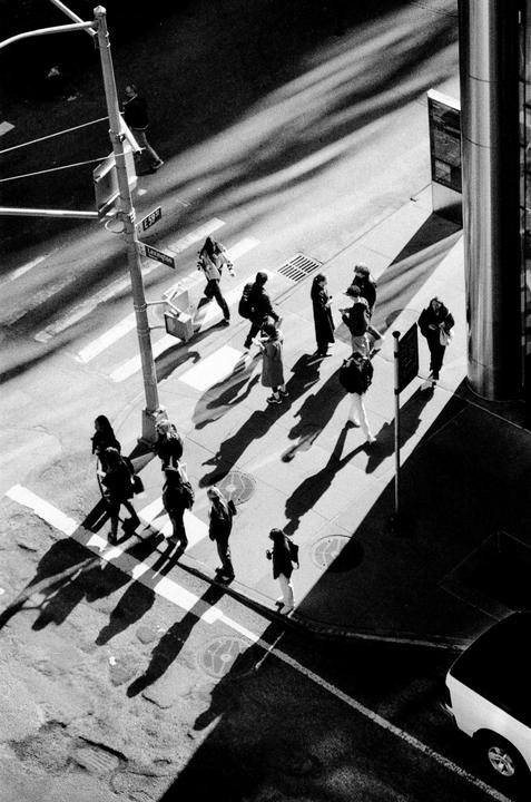 A high-angle view shows pedestrians crossing a street with long shadows cast across the pavement.