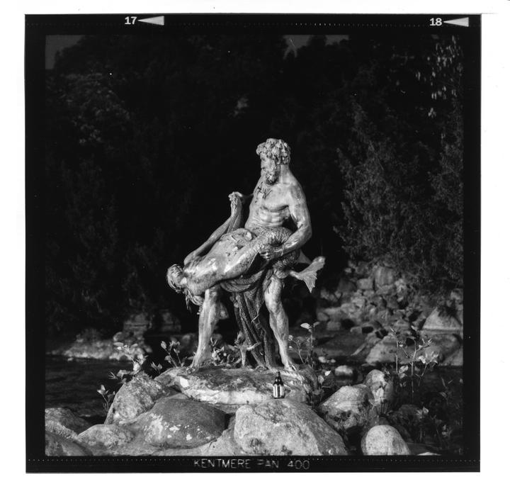 A statue of a man holding a mermaid stands on rocks with dark foliage in the background.
